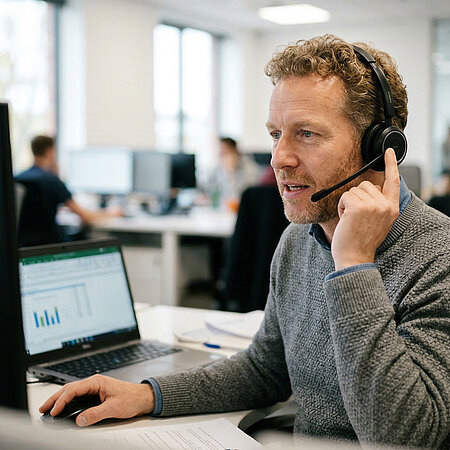 Mitarbeiter mit Headset arbeitet im Büro am Computer und spricht mit einem Kunden.