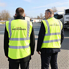 Zwei WestCrown-Mitarbeiter in Warnwesten gehen über das Werksgelände mit Lkw im Hintergrund.