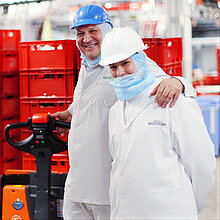 Zwei WestCrown-Mitarbeitende in Hygienekleidung stehen lächelnd im Produktionsbereich vor roten Transportkisten.