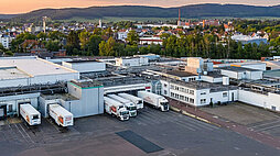 Luftaufnahme des WestCrown-Werks mit Produktionsgebäuden, Laderampen und Lkw vor einer Kleinstadt und bewaldeten Hügeln.
