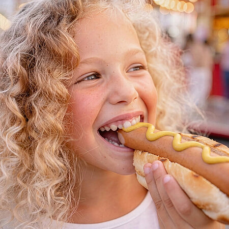 Kind isst einen Hotdog mit Senf und lächelt dabei.