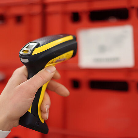 Hand scannt das Etikett einer roten Transportkiste mit einem Barcodescanner.