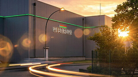 WestCrown-Gebäude im Abendlicht mit beleuchtetem Logo, grüner Fassadenlinie und Lichtspuren auf der Straße.