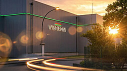 WestCrown-Gebäude im Abendlicht mit beleuchtetem Logo, grüner Fassadenlinie und Lichtspuren auf der Straße.